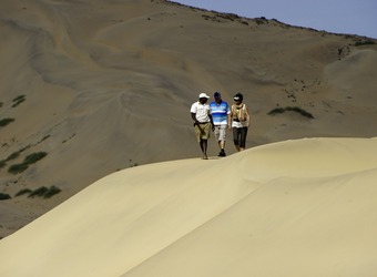 Three safari guests walk along the ridge of a massive beige colored sand dune in the Hartmann Valley, Namibia