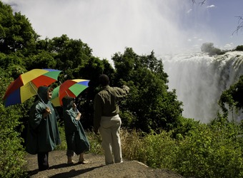 A guide informs on Victoria Falls to two tourists with vibrantly promary colored umbrellas wearing ponchos in Zimbabwe.